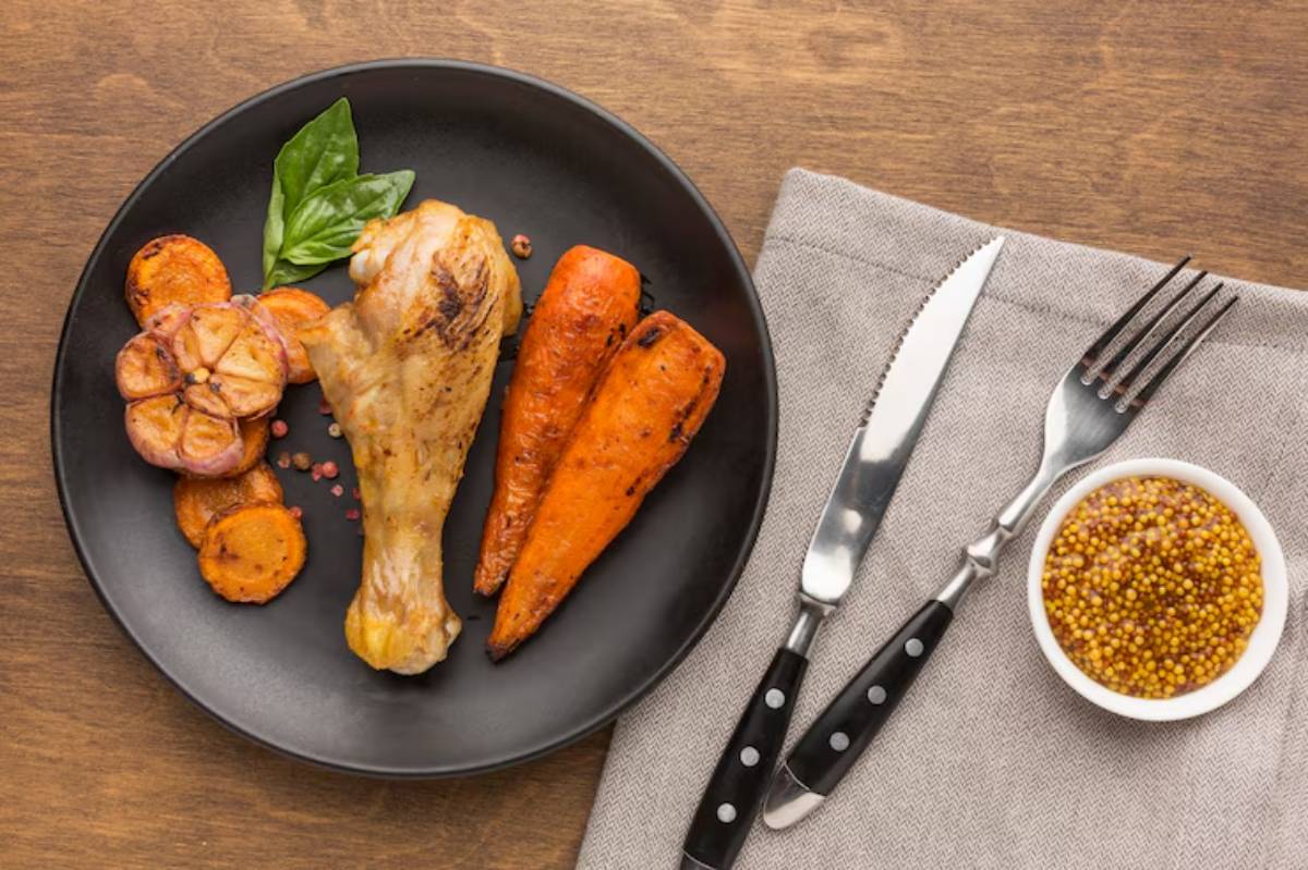 A black plate with roasted chicken leg, carrots, garlic, and basil, accompanied by mustard in a bowl and cutlery on a linen napkin.