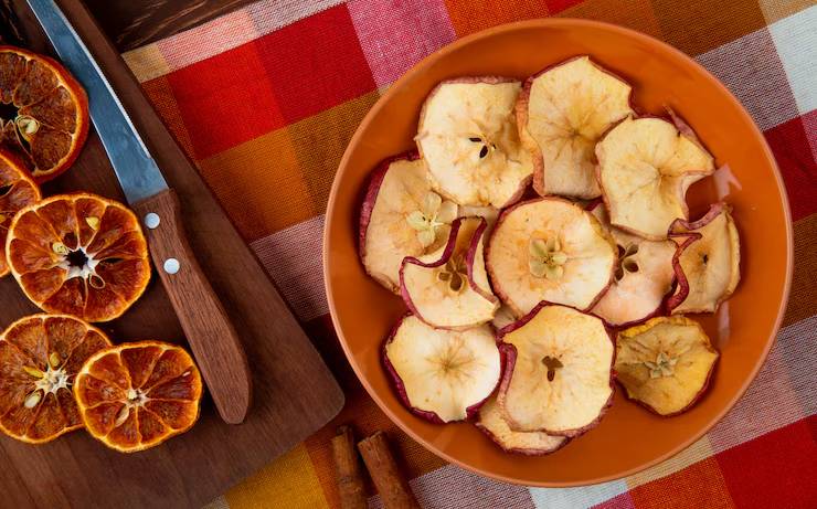 A bowl of dried apple slices surrounded by dehydrated orange slices, a knife, and cinnamon sticks on a colorful plaid tablecloth.
