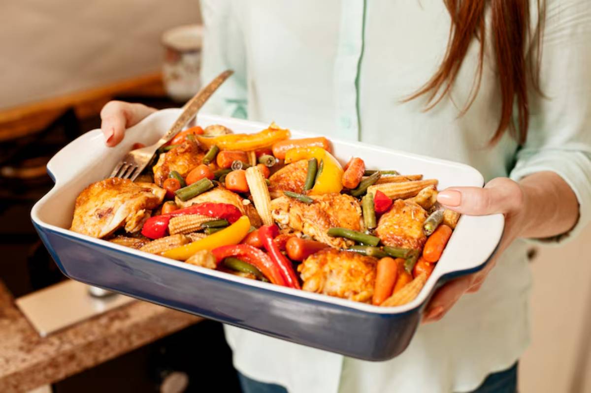 A woman holds a baking dish filled with roasted chicken and colorful vegetables, ready to serve a delicious meal.
