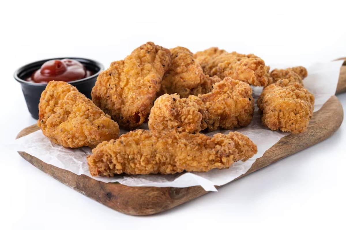 A plate of crispy fried chicken pieces served with a small bowl of ketchup on a wooden board lined with parchment paper.