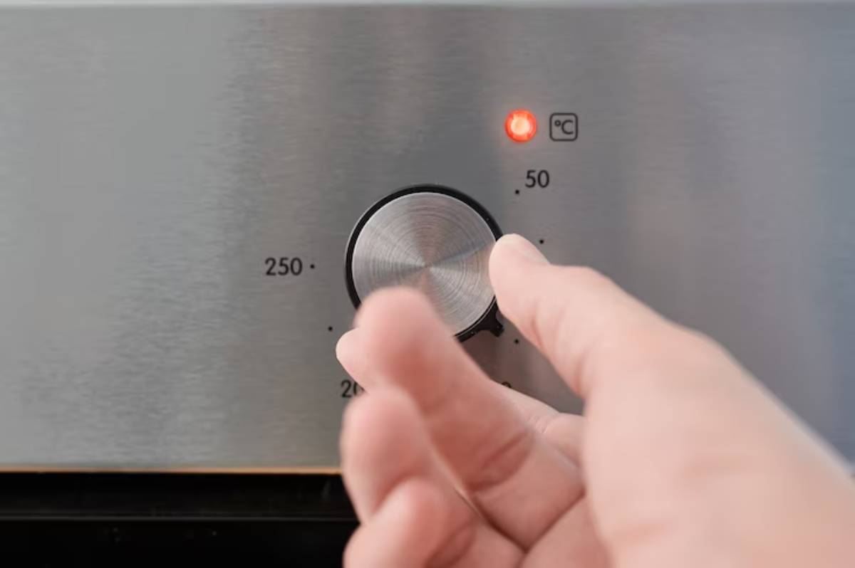 A hand adjusts a temperature knob on a stainless steel appliance, with a red indicator light glowing above it.