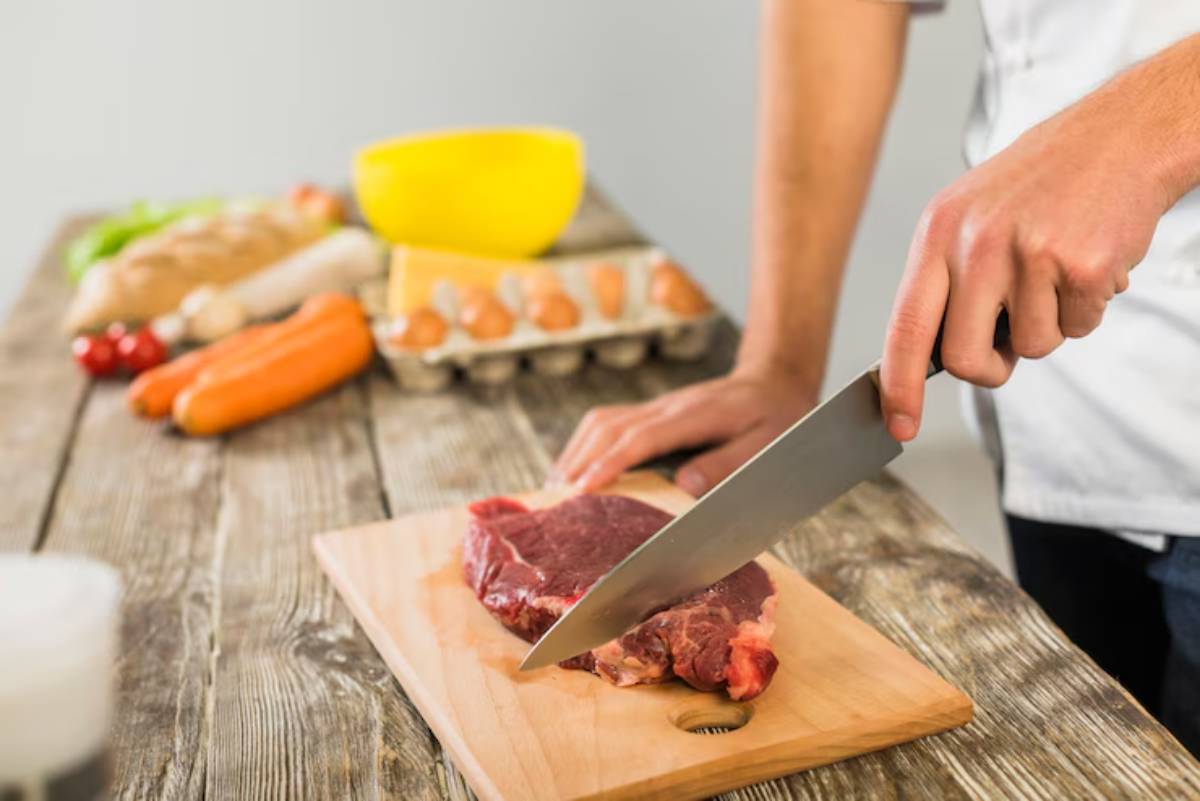 A person slices a steak on a wooden cutting board, surrounded by fresh vegetables, eggs, and a yellow bowl in the kitchen.