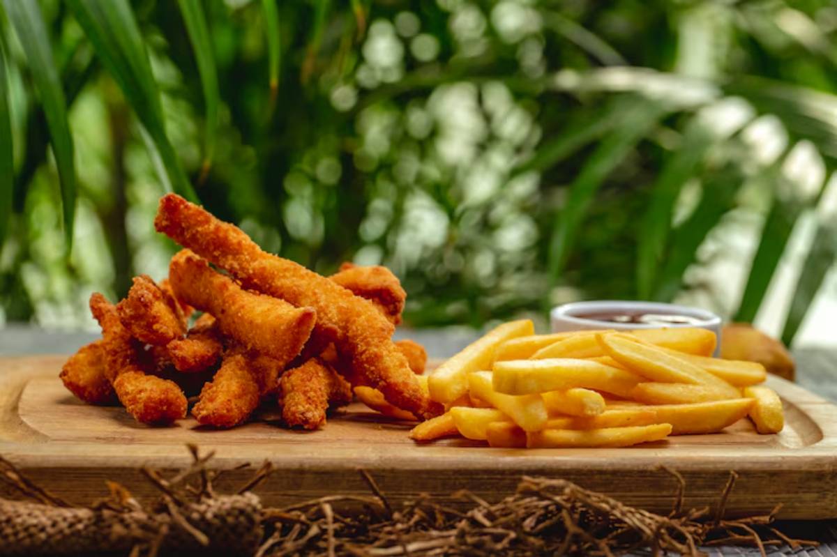 Crispy fried chicken strips and golden French fries served on a wooden platter, garnished with a side of dipping sauce and a lush green backdrop.