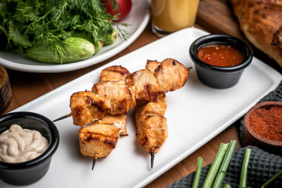 Grilled chicken skewers on a plate, accompanied by dipping sauces and fresh herbs, with a glass of juice and bread in the background.