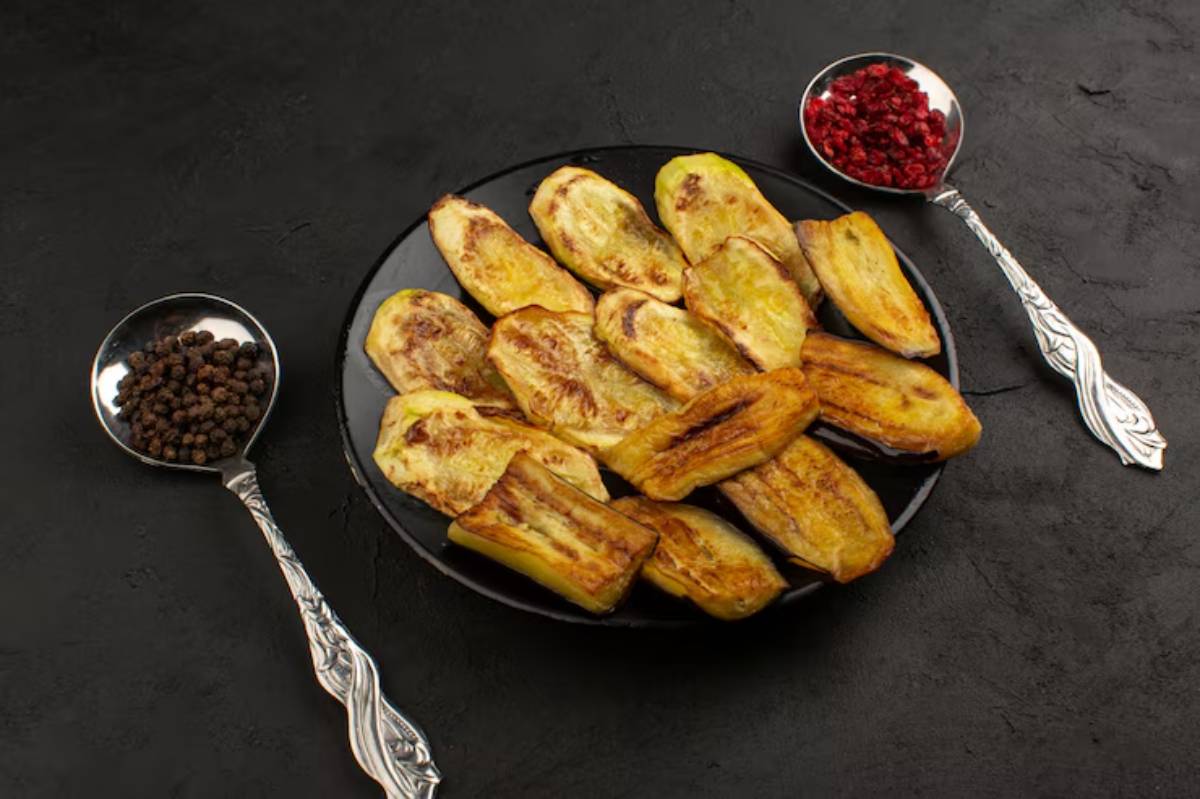 A black plate with golden-brown fried eggplant slices, accompanied by silver spoons of black pepper and dried red fruits.
