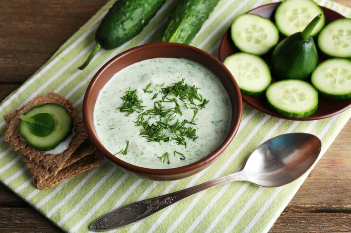 A bowl of creamy cucumber soup garnished with fresh herbs, accompanied by slices of cucumber and crispbread on a striped cloth background.
