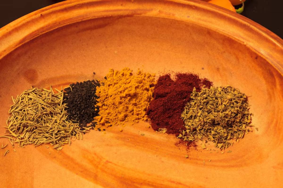 A rustic plate featuring five vibrant piles of spices: cumin seeds, black seed, turmeric, paprika, and dried herbs.