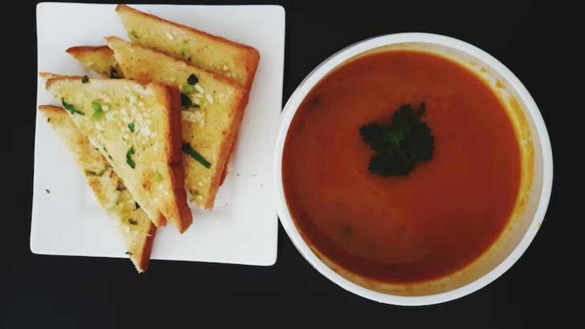 A bowl of rich tomato soup garnished with cilantro, accompanied by crispy garlic bread slices on a white plate.