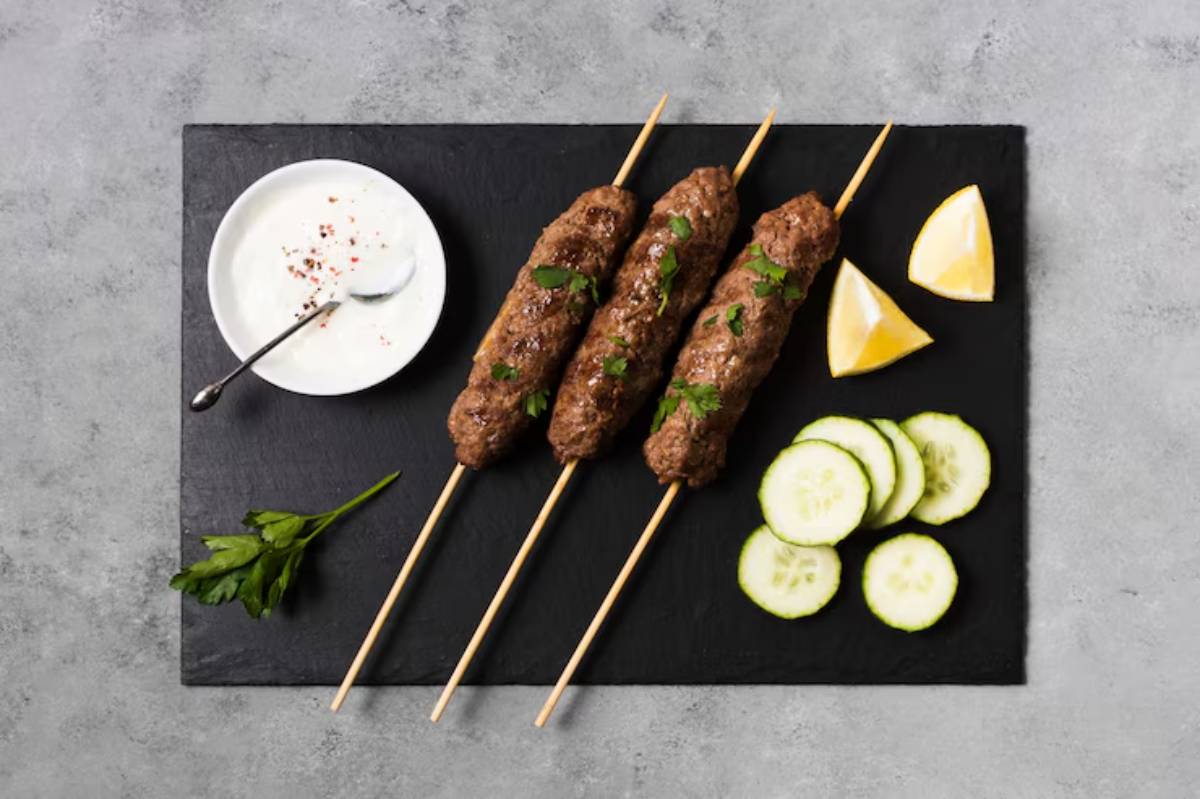 Three skewers of grilled meat topped with parsley, served with cucumber slices, lemon wedges, and yogurt sauce on a black slate board.
