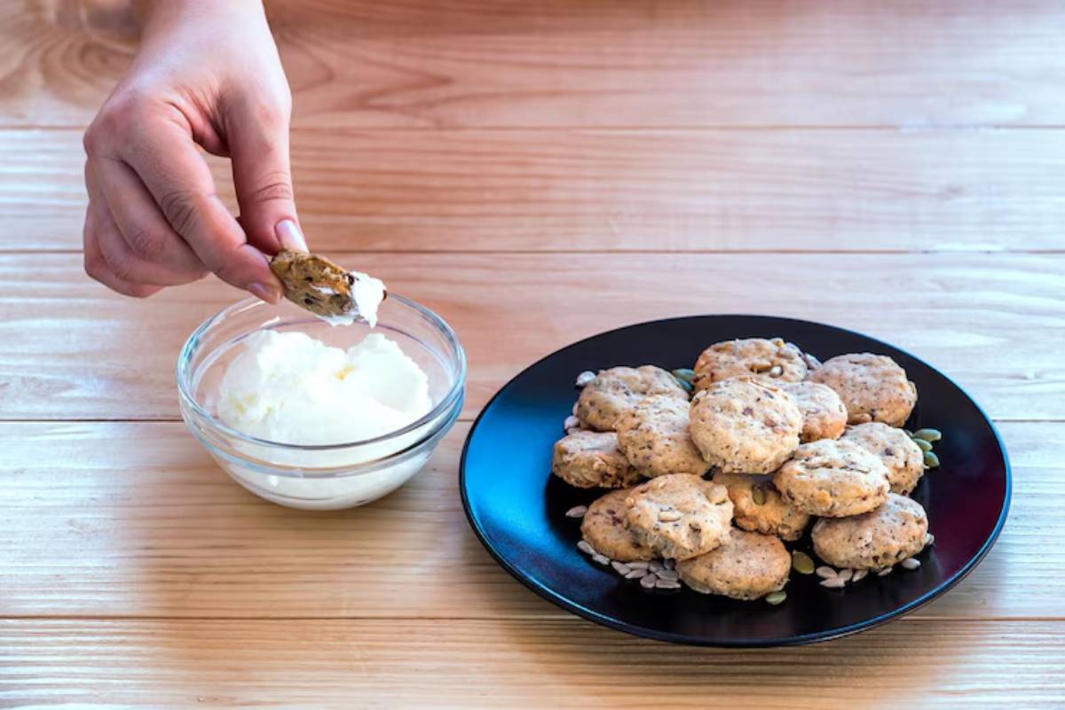 A hand dips a cookie into a bowl of creamy dip, with a plate of cookies on a wooden table in the background.