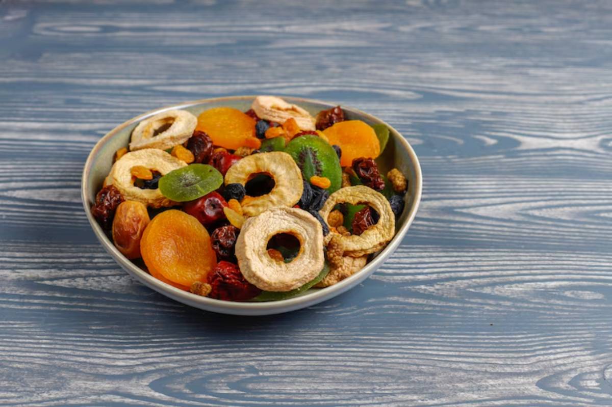 A bowl of colorful dried fruits, including apple rings, apricots, and raisins, arranged beautifully on a blue wooden surface.