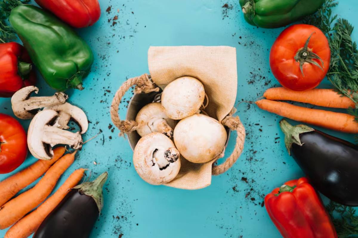 Basket of white mushrooms surrounded by vibrant vegetables: carrots, red and green bell peppers, tomatoes, eggplant, and fresh herbs.