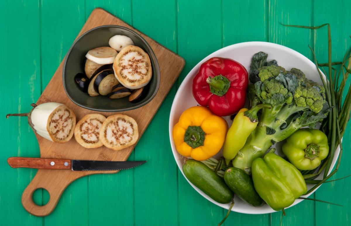 A wooden cutting board with sliced eggplant and a knife beside a bowl of colorful vegetables, including peppers, broccoli, and cucumbers.