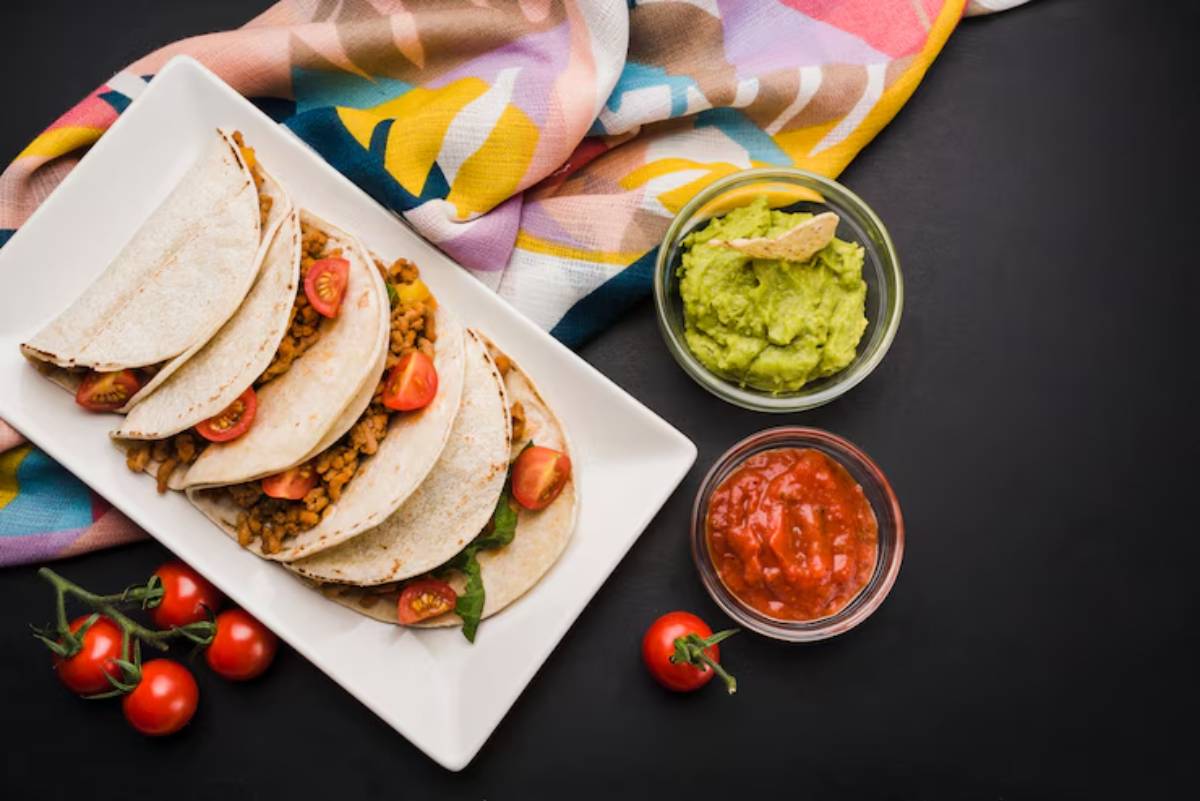 A plate of tacos filled with seasoned meat and cherry tomatoes, accompanied by guacamole and salsa, beside fresh cherry tomatoes.