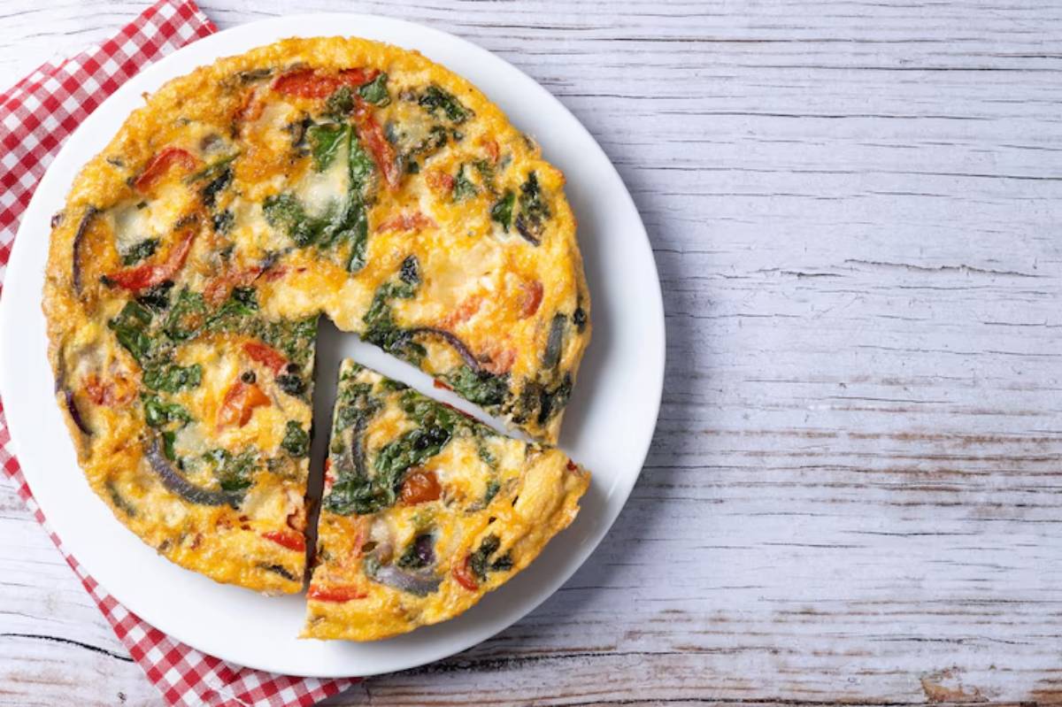 A vegetable frittata with spinach, red peppers, and onions, partially sliced, on a white plate with a red checkered napkin.