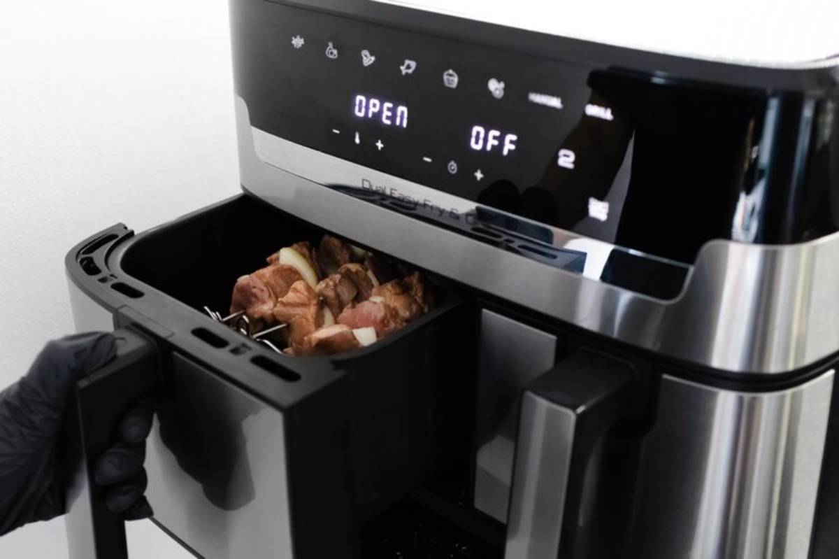 A hand in a black glove opens an air fryer, revealing marinated meat inside, with digital controls displayed on the front.