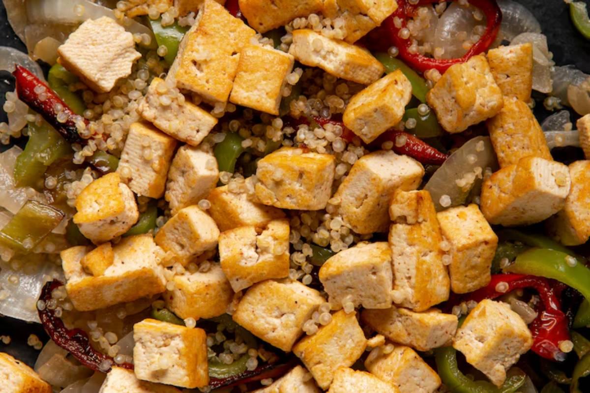 A close-up of stir-fried quinoa with cubed tofu, bell peppers, and red chili peppers on a dark surface.
