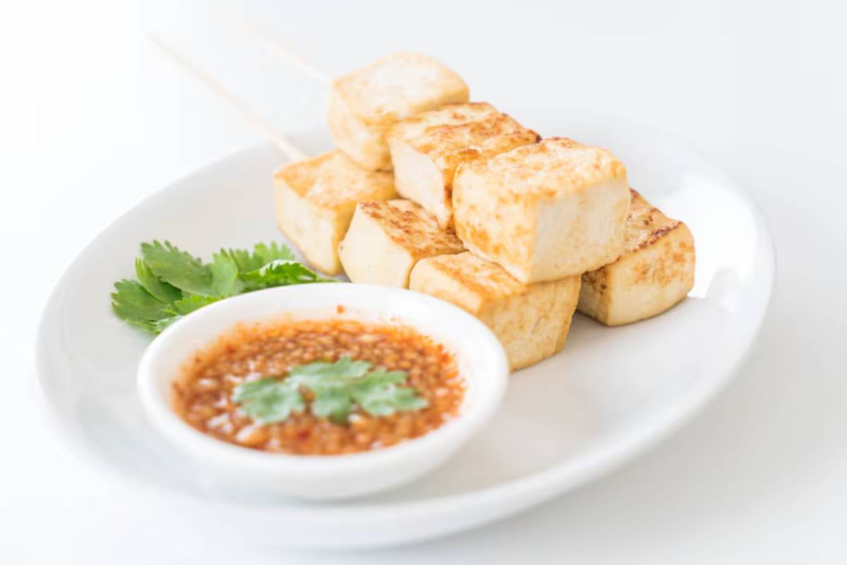Grilled tofu skewers served with a spicy dipping sauce and garnished with fresh cilantro on a white plate.