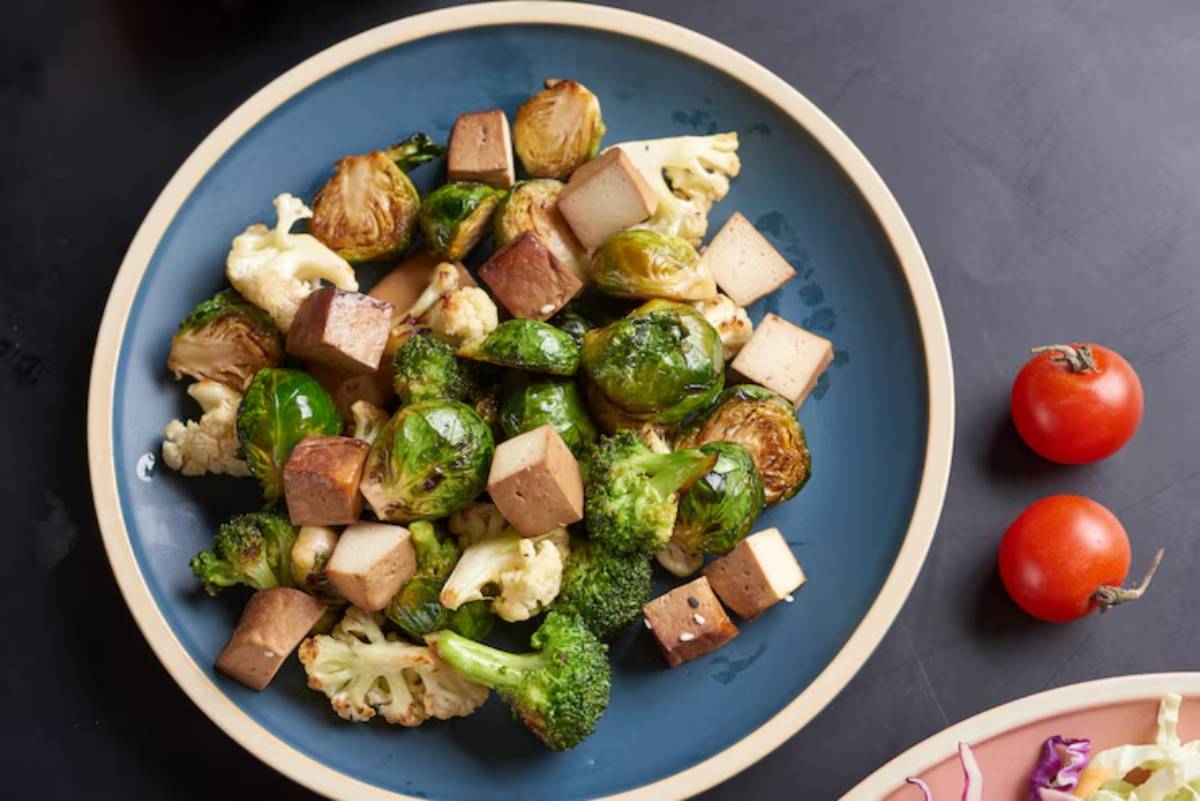 A colorful plate of roasted Brussels sprouts, broccoli, cauliflower, and tofu cubes, accompanied by two cherry tomatoes.