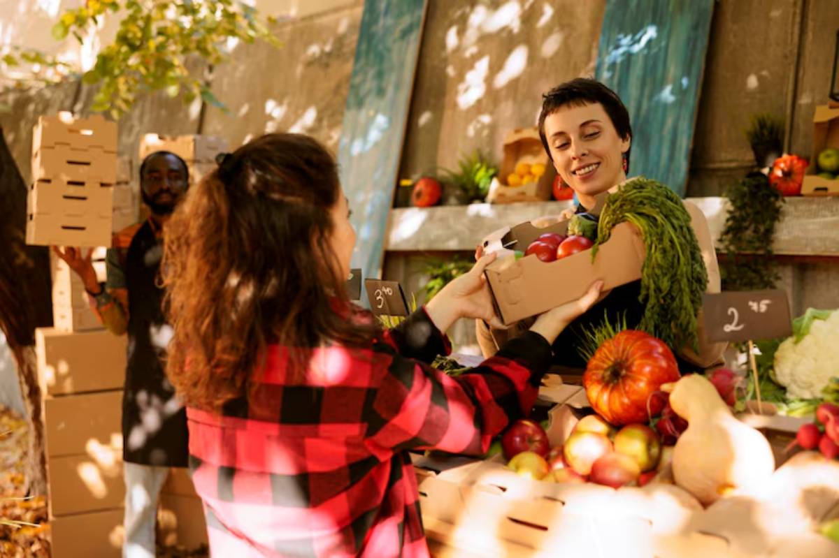 A woman in a black and red plaid shirt hands a box of produce to a vendor at a vibrant outdoor market filled with fresh vegetables and fruits.