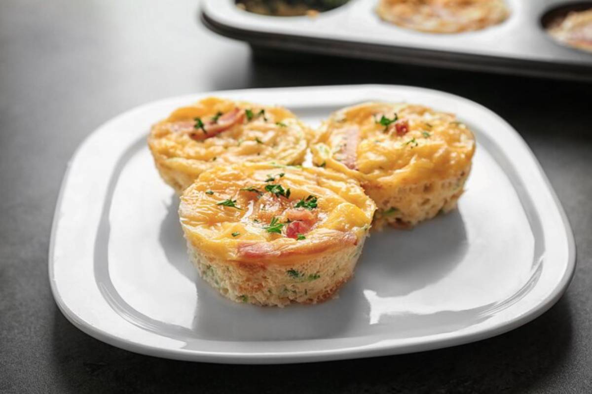 Three savory egg muffins with cheese, ham, and herbs, served on a white plate, with additional muffins in a muffin tray in the background.