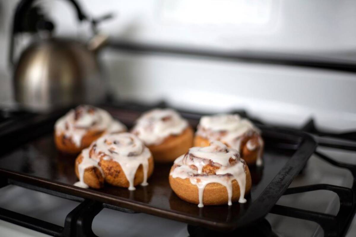Freshly baked cinnamon rolls topped with creamy icing, resting on a dark baking tray beside a kettle on a gas stove.