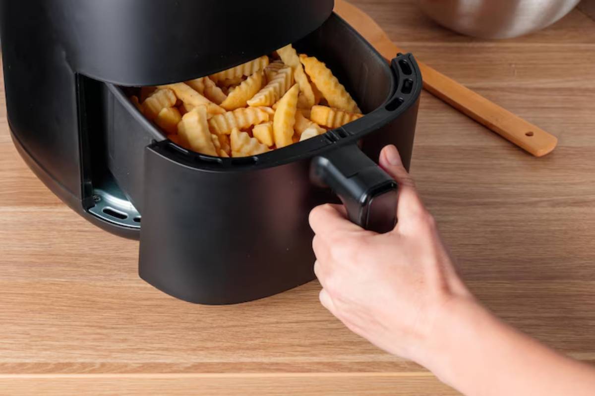 A hand grasps the handle of an open black air fryer filled with golden crinkle-cut fries on a wooden countertop.