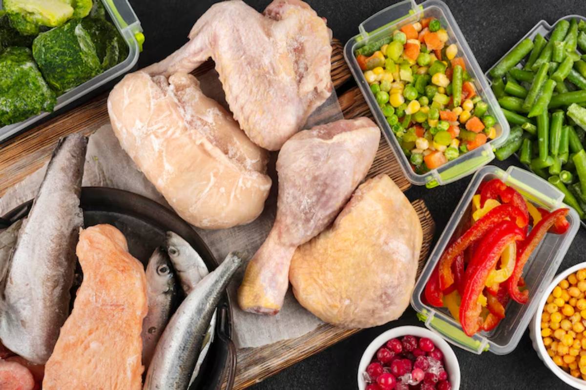 A variety of frozen foods including chicken pieces, fish, vegetables, and berries arranged on a wooden surface.