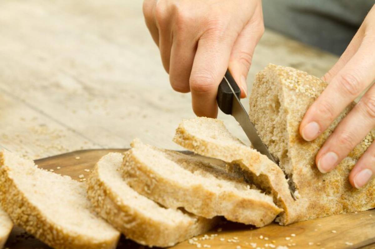 A hand holds a knife, slicing through fresh whole grain bread on a wooden cutting board.
