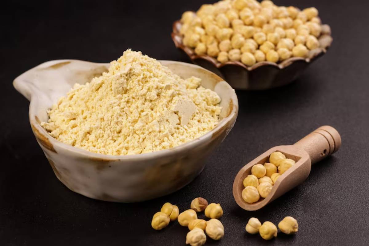 A bowl of chickpea flour, a dish of whole chickpeas, and a wooden scoop filled with chickpeas on a dark background.
