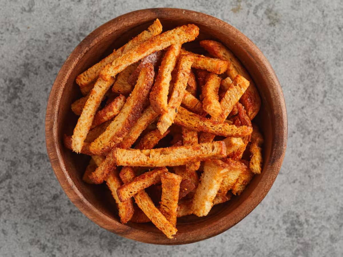 A wooden bowl filled with golden-brown, crispy breadsticks, dusted with seasoning, set against a gray stone background.