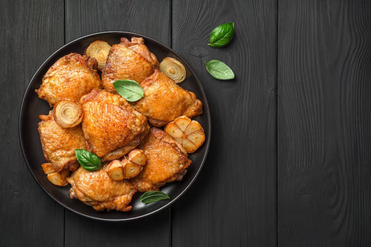 A black plate of golden-brown roasted chicken thighs garnished with fresh basil and roasted garlic on a dark wooden table.