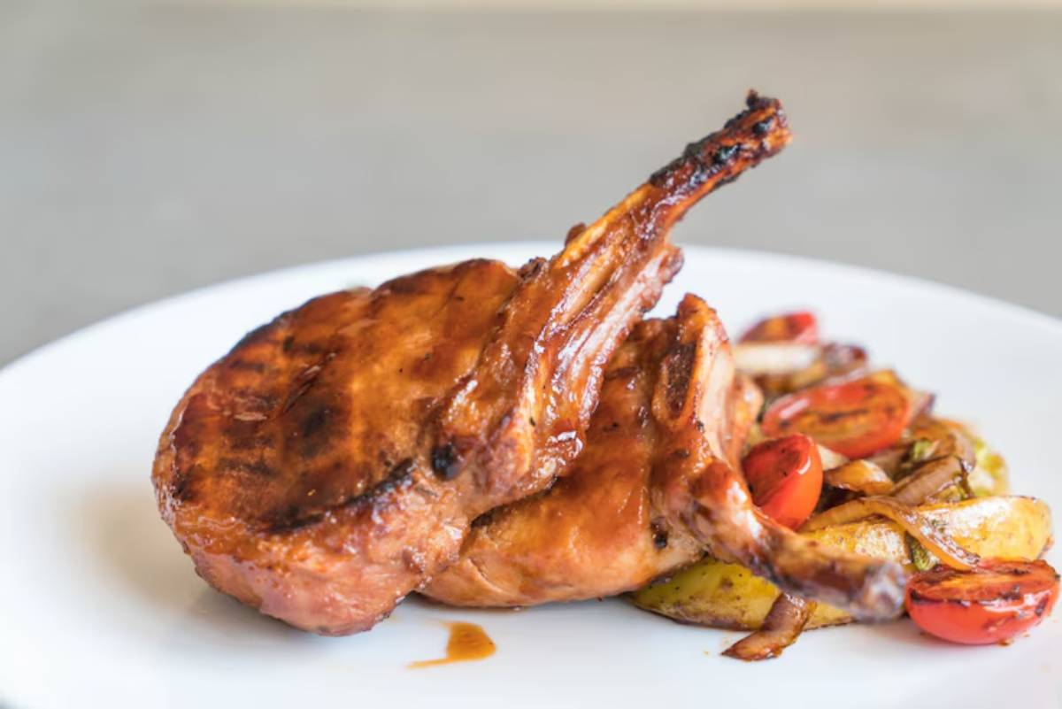 Grilled pork chop with a caramelized glaze, served alongside sautéed onions and colorful cherry tomatoes on a white plate.