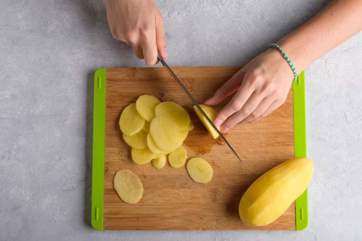 A hand slices peeled potatoes on a wooden cutting board with a green edge, surrounded by freshly cut potato slices.