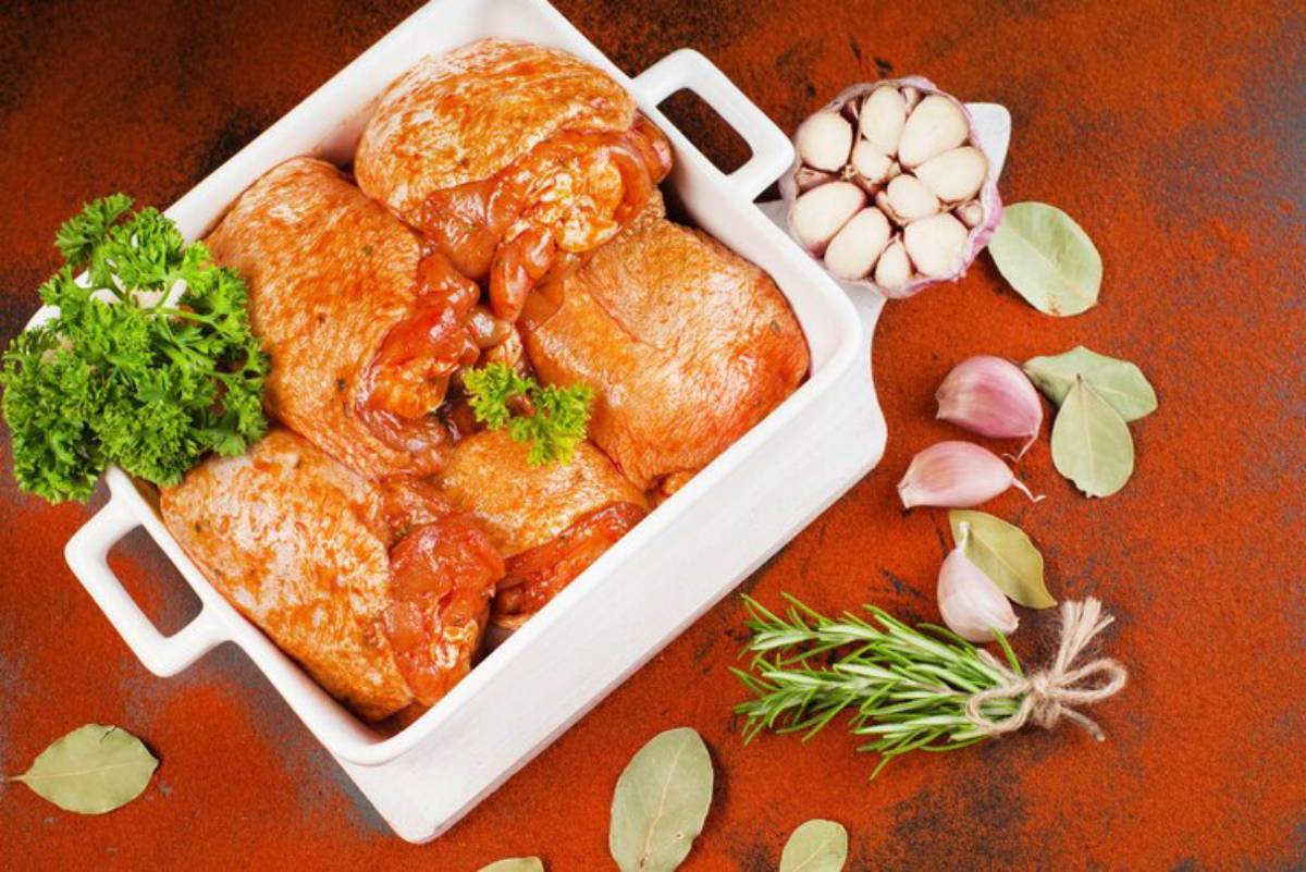A white dish holds marinated chicken thighs, garnished with parsley, surrounded by garlic, bay leaves, and fresh rosemary on a rustic surface.