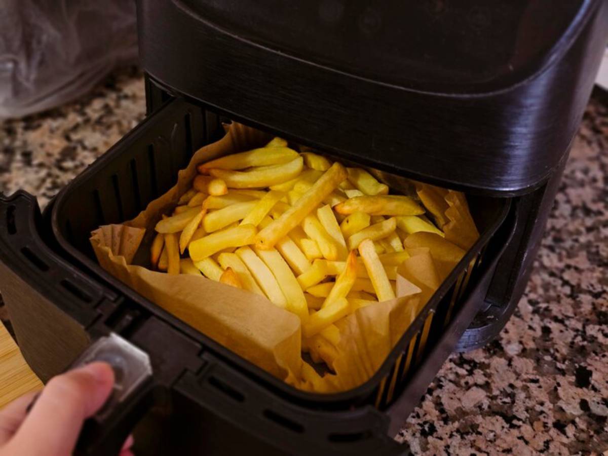 A hand holding the basket of an air fryer filled with golden, crispy French fries lined with parchment paper.