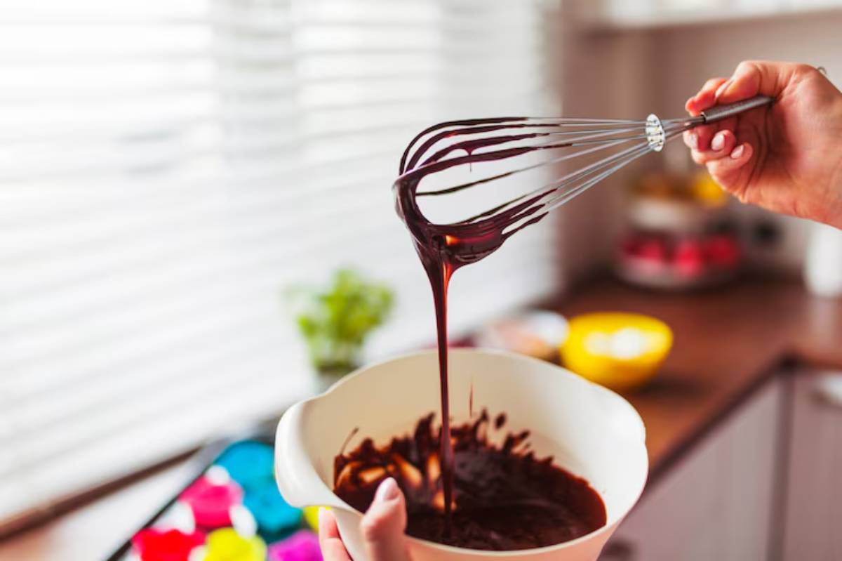A hand holding a whisk over a bowl of dark chocolate batter in a bright kitchen, with colorful bowls in the background.