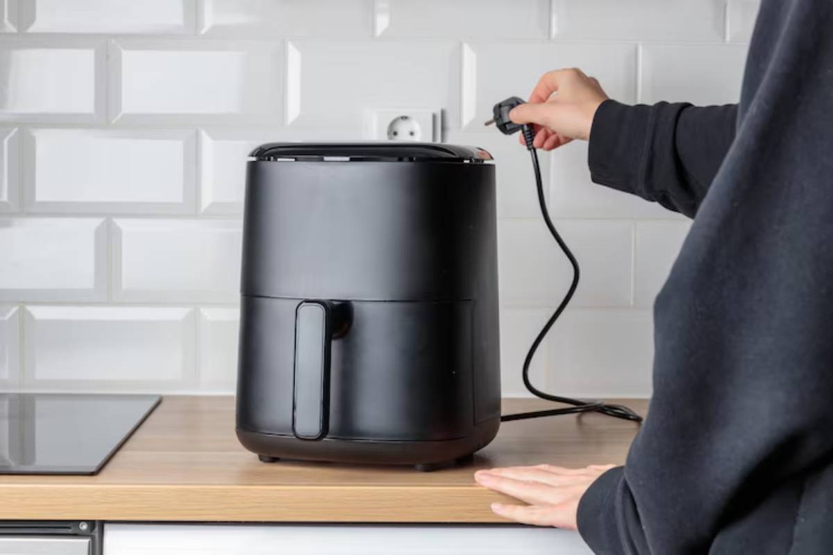 A person plugs in a black air fryer on a kitchen countertop against a glossy white tiled wall.