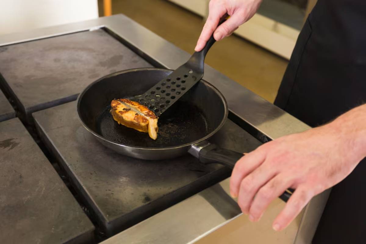 A chef's hand uses a spatula to flip a piece of chicken in a frying pan on a stovetop. The chicken has a golden-brown crust.