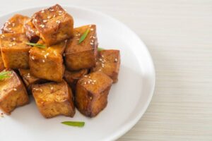 A plate of golden-brown, caramelized tofu cubes garnished with sesame seeds and green onions, set against a light background.