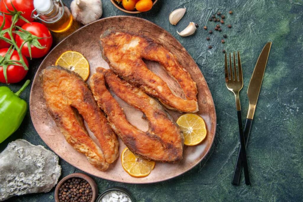 Three golden-brown fried fish fillets on a platter, surrounded by sliced lemons, tomatoes, garlic, peppers, and spices.