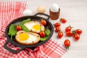 A cast iron skillet with two eggs in toasted bread, surrounded by spinach and cherry tomatoes, on a red checkered cloth. Eggs in the background.
