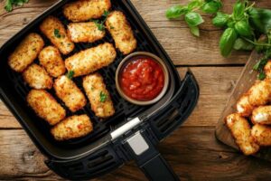 Crispy, golden-brown cheese sticks in an air fryer basket, accompanied by a bowl of marinara sauce on a rustic wooden table.