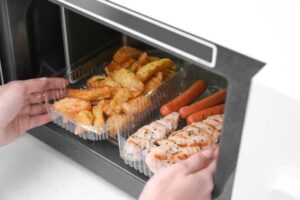 A person placing a clear container of golden fries into a microwave next to sausages and grilled chicken fillets.