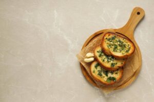 Three slices of garlic bread topped with parsley, served on a wooden board with two garlic cloves, set against a light marble background.