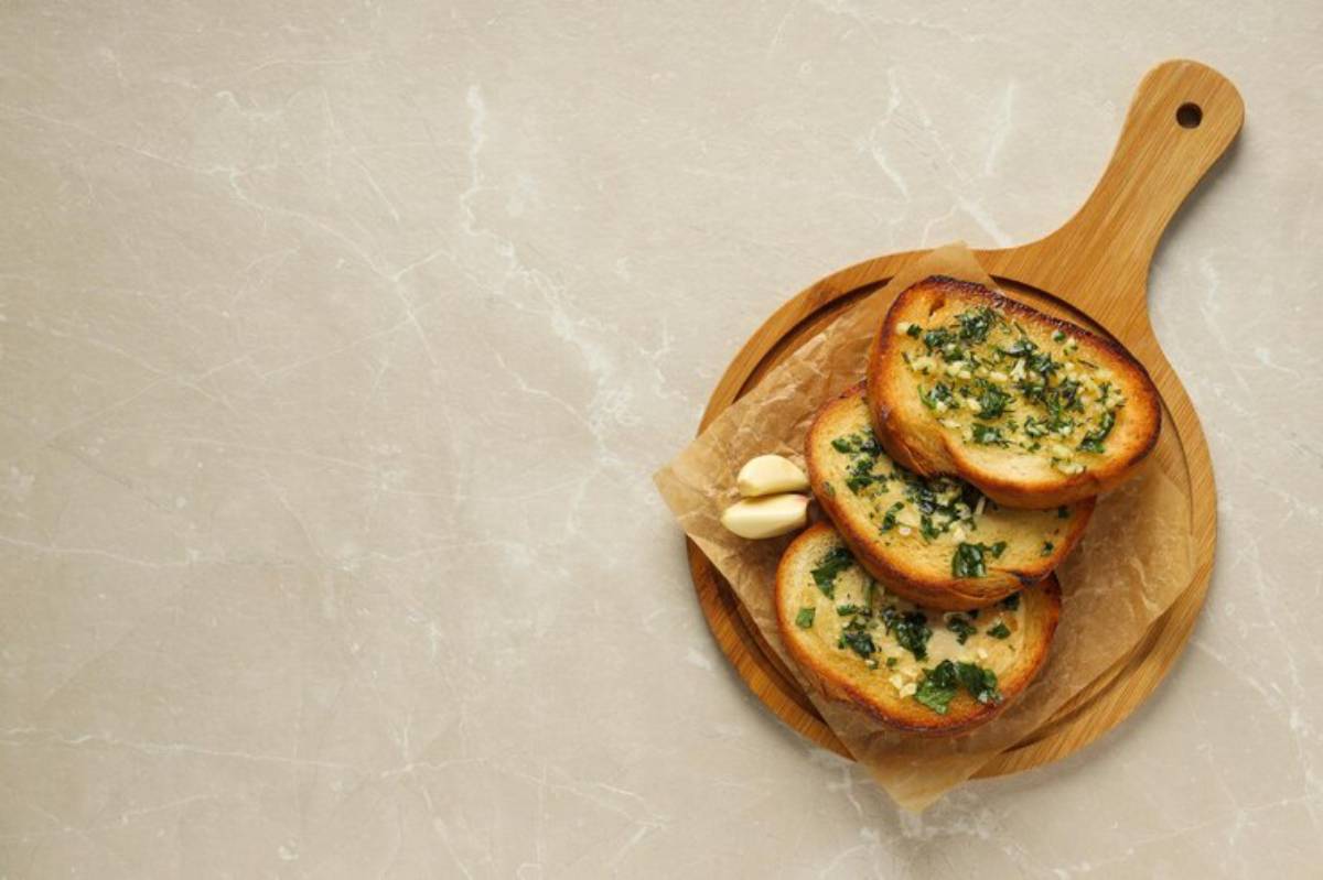 Three slices of garlic bread topped with parsley, served on a wooden board with two garlic cloves, set against a light marble background.