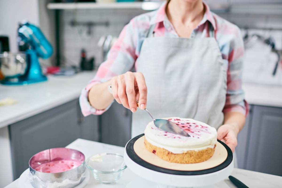 A woman in a plaid shirt and apron smooths frosting on a layered cake, with baking tools and ingredients visible in a bright kitchen.