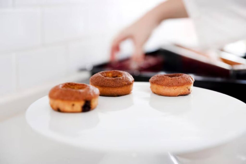 Three freshly baked donuts on a white plate, with a blurred hand reaching for the baking tray in the background.