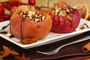 Two baked apples stuffed with nuts and drizzled with chocolate sauce, served on a white plate with spices and festive decorations.