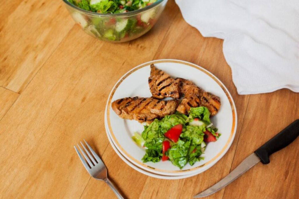 A black plate of golden-brown roasted chicken thighs garnished with fresh basil and roasted garlic on a dark wooden table.